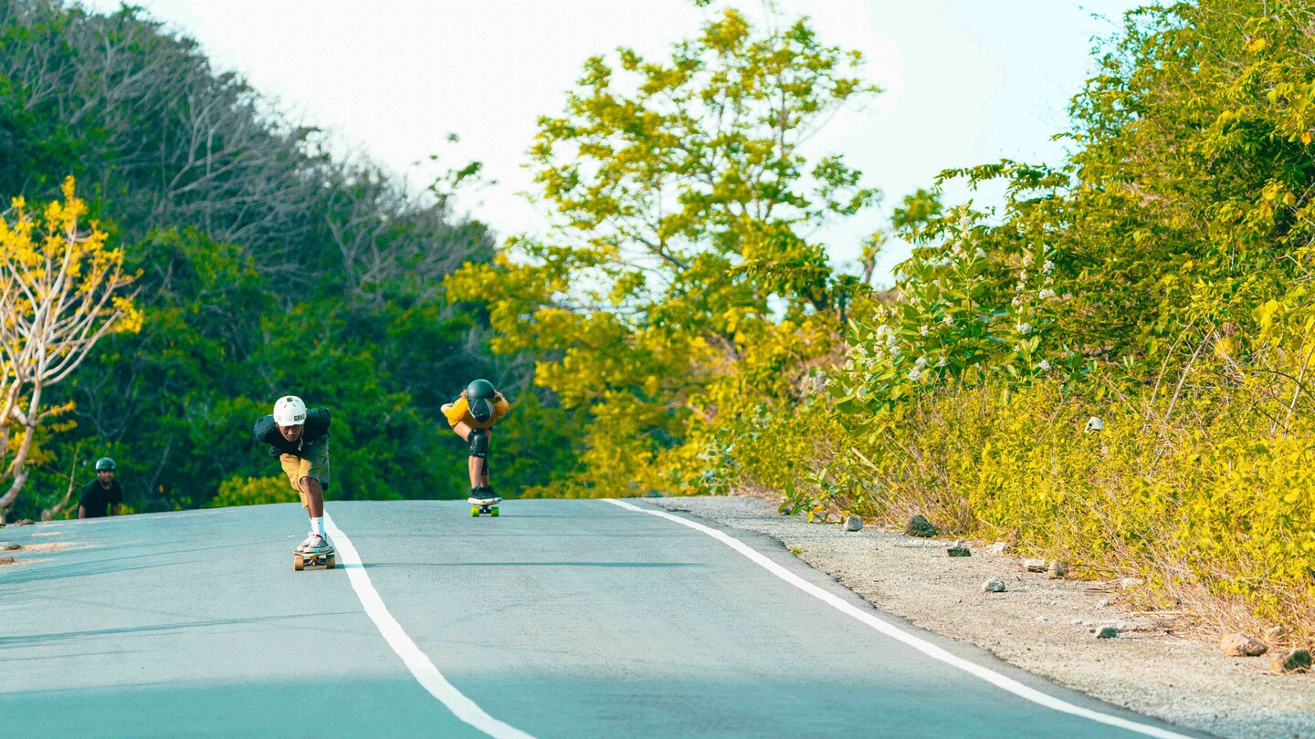 Mehrere Downhill-Skateboarder, darunter Anna Pixner mit gelbem T-Shirt und schwarzem Helm, fahren eine ländliche, leicht abschüssige Strasse hinunter. Die Szene ist von üppiger Vegetation und Bäumen umgeben.