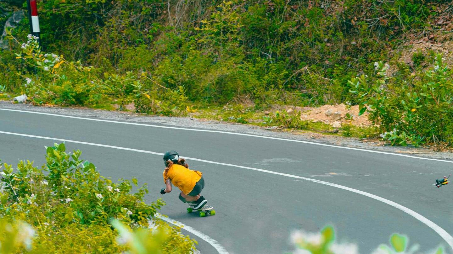 Anna Pixner, eine Downhill-Skateboarderin, fährt in gebückter Position mit einem Skateboard eine enge Kurve auf einer ländlichen Strasse hinunter. Sie trägt einen schwarzen Helm, ein gelbes T-Shirt und Schutzausrüstung. Die Umgebung ist von dichter Vegetation umgeben.