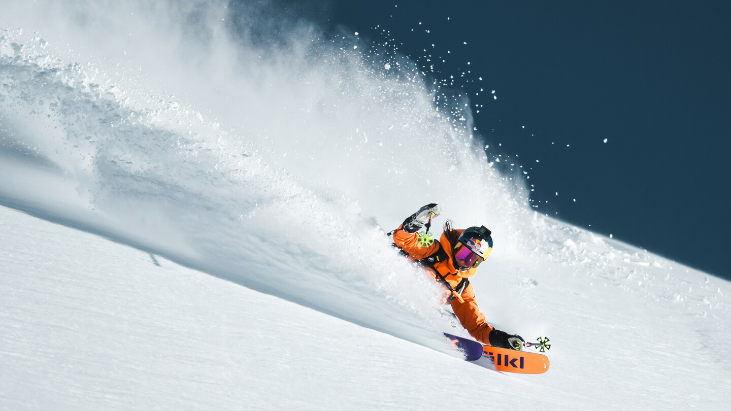 Skifahrerin in orangefarbener Jacke fährt dynamisch durch tiefen Pulverschnee, aufwirbelnder Schnee im Hintergrund.