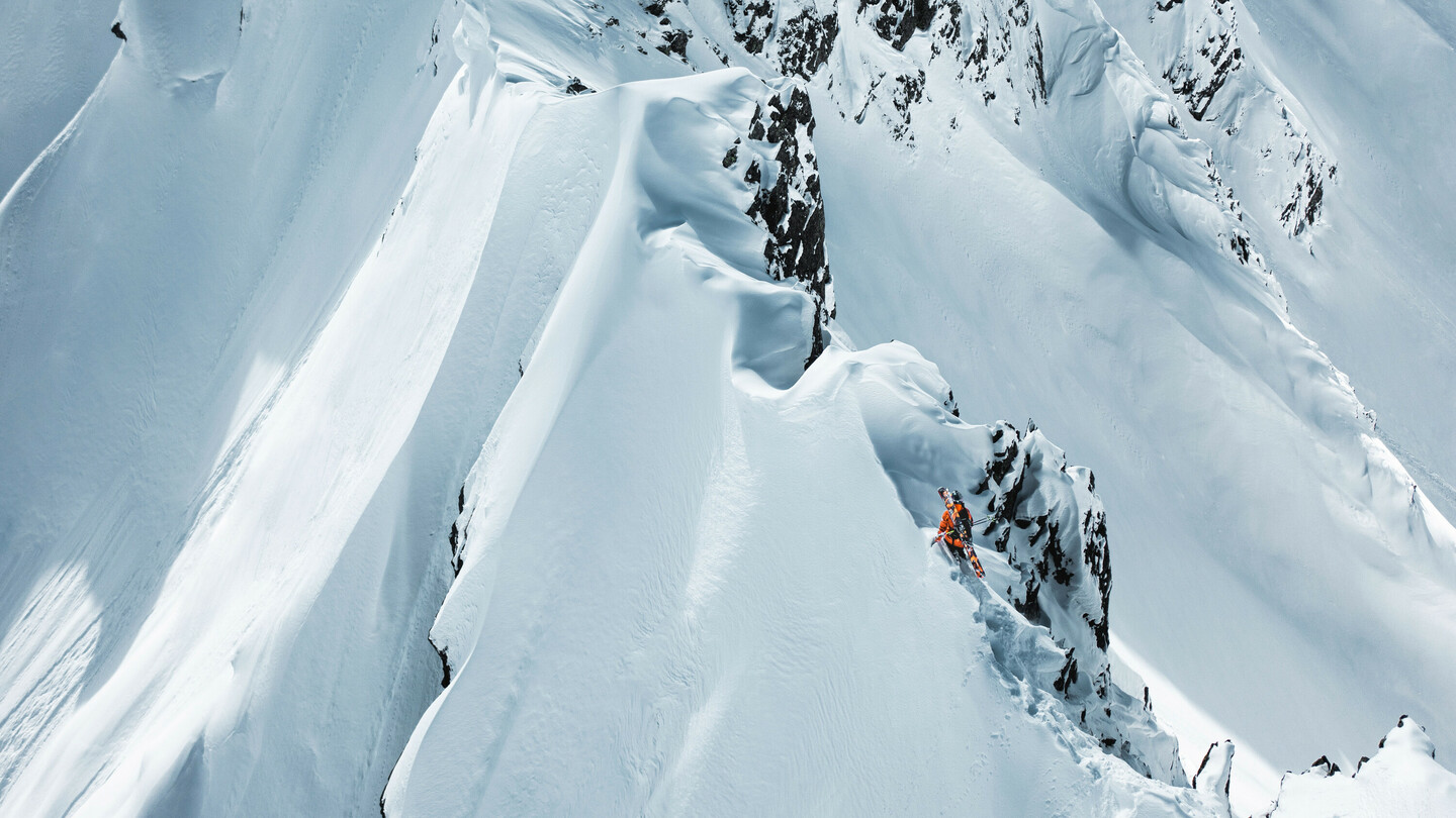 Skifahrer in orangefarbener Jacke navigiert vorsichtig entlang einer schmalen, steilen Schneekante in einer dramatischen Berglandschaft.