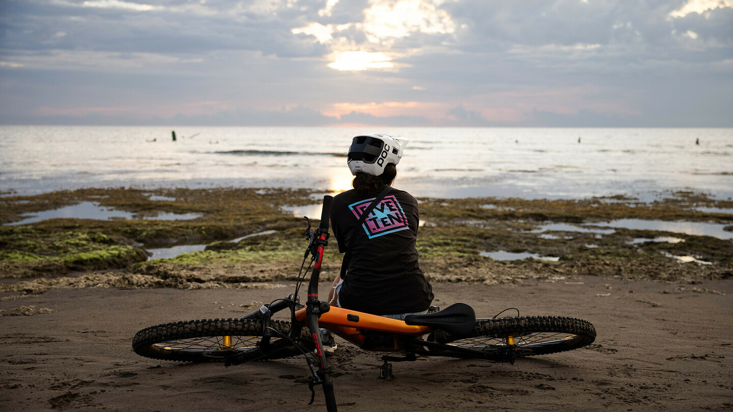 Eine Person mit Helm sitzt am Strand neben einem Mountainbike und blickt auf das Meer, während die Sonne hinter den Wolken untergeht. Die Stimmung ist ruhig und entspannt.