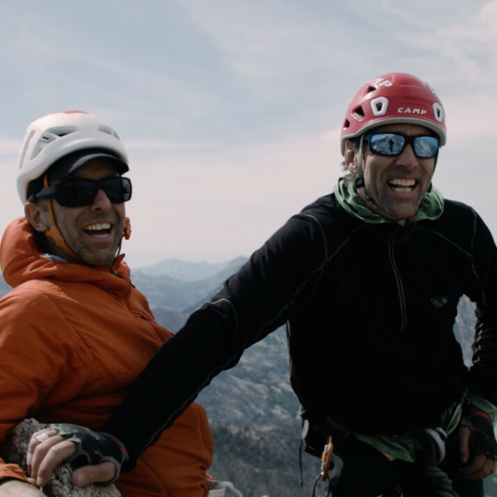 On the left stands Timmy O’Neill wearing an orange jacket and helmet, smiling with sunglasses on. To his right stands blind climber Erik Weihenmayer in a black jacket and red helmet. Both men are on a mountain summit, appearing happy and enjoying the vast mountainous landscape in the background. The image captures the spirit of climbing, adventure, and determination.