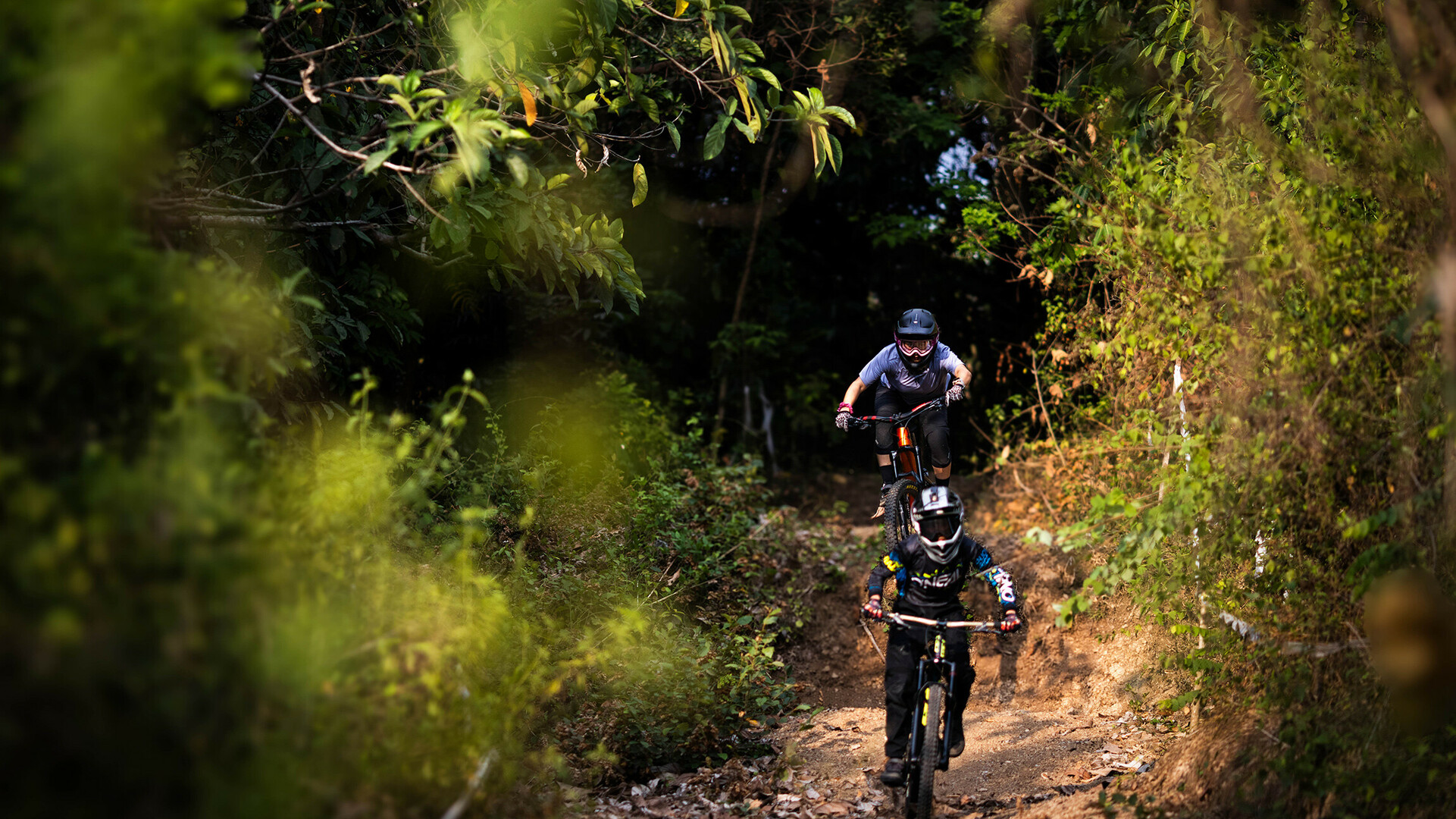 Zwei Mountainbiker fahren einen schmalen, von Bäumen umgebenen Pfad hinunter. Der vordere Fahrer trägt eine schwarze Ausrüstung mit einem Helm, während der hintere Fahrer eine graue Kleidung und eine Schutzbrille trägt.